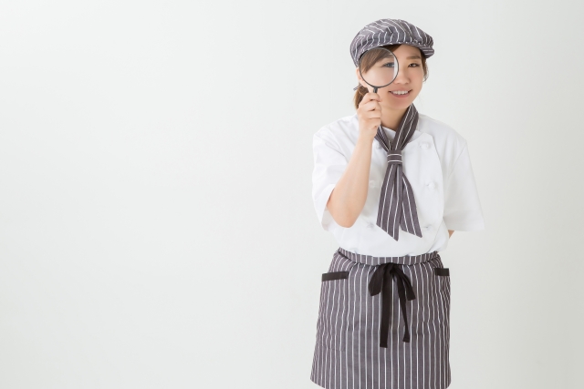 コックコートを着た女性が虫眼鏡を覗いている様子。アルバイト探しをイメージ。