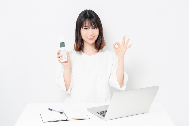 ノートパソコンの前で電話を持ってOKサインをする女性